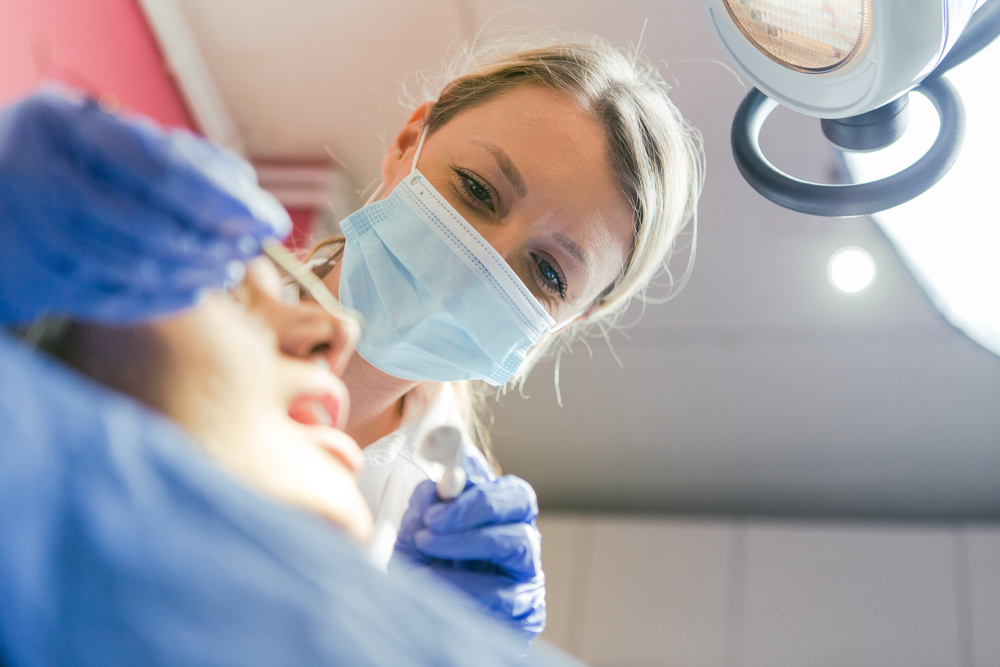 A healthcare professional wearing a mask and gloves examines a patient lying down, likely in a medical or dental setting, under a bright examination light.