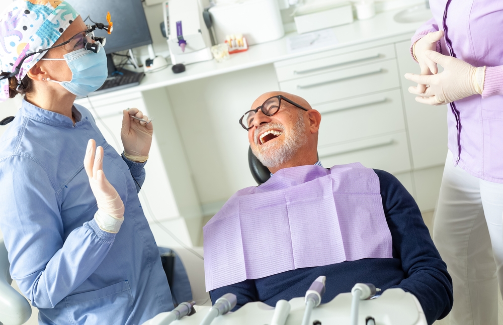 An older man with glasses sits in a dental chair, smiling and laughing, as a dental professional in scrubs and gloves talks to him in a bright, modern dental office. Dental tools are visible in the foreground.