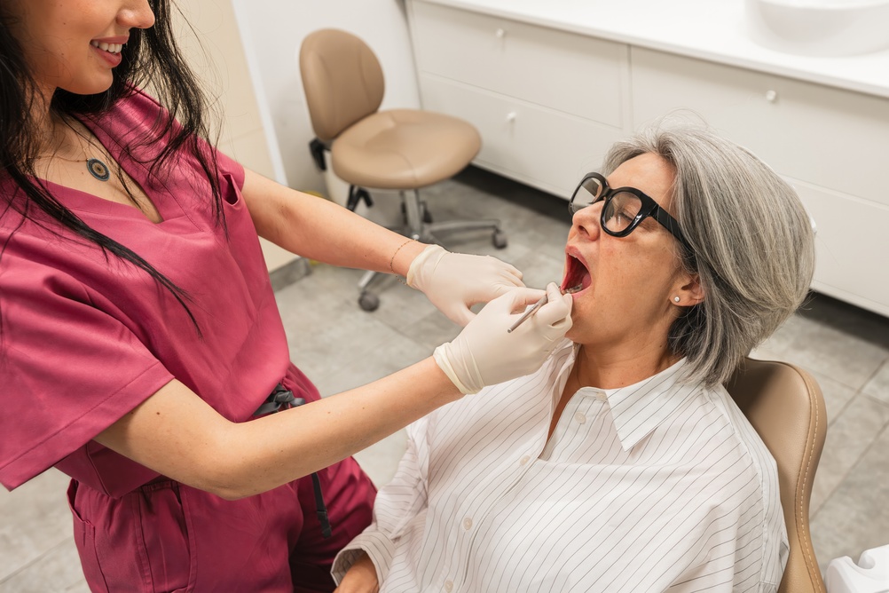A dentist in pink scrubs and gloves examines the open mouth of an older woman with gray hair and black glasses, who is seated in a dental chair in a clinic room.