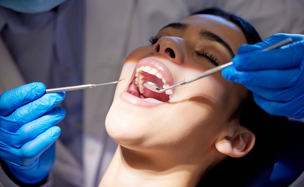 A person reclines in a dental chair with their mouth open while a dentist wearing blue gloves uses dental instruments to check their teeth and gums under bright lighting.