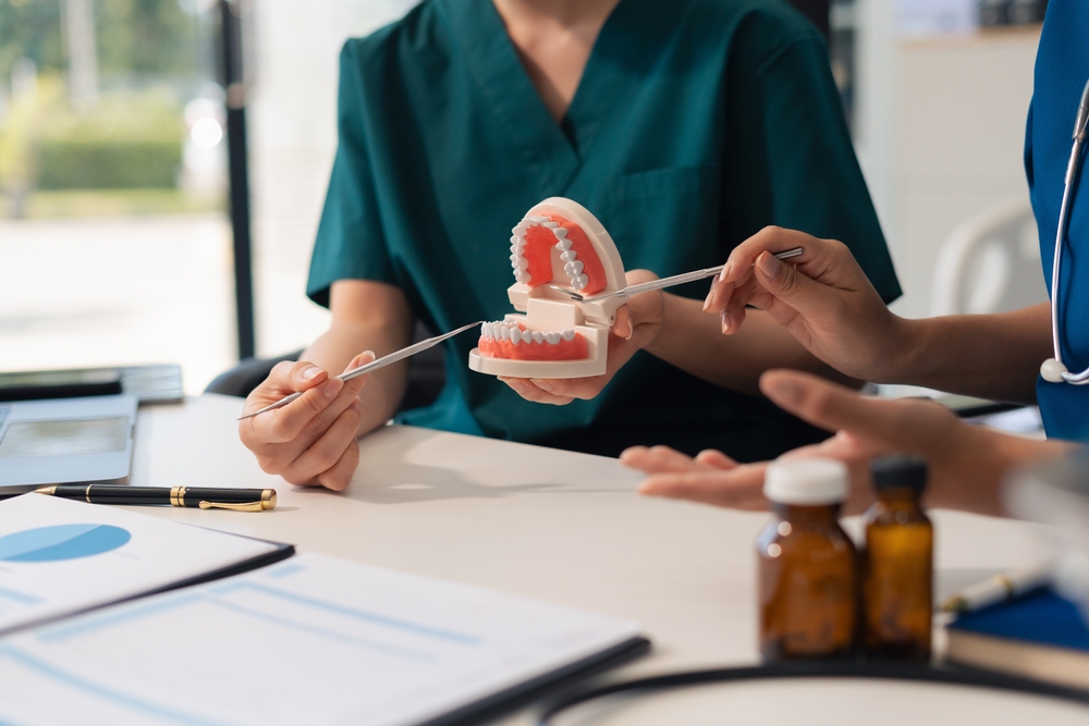 Two people sit at a table; one in medical scrubs points at a dental model with a pen, explaining oral hygiene. The other person's hands are visible. Medicine bottles and paperwork rest on the table.