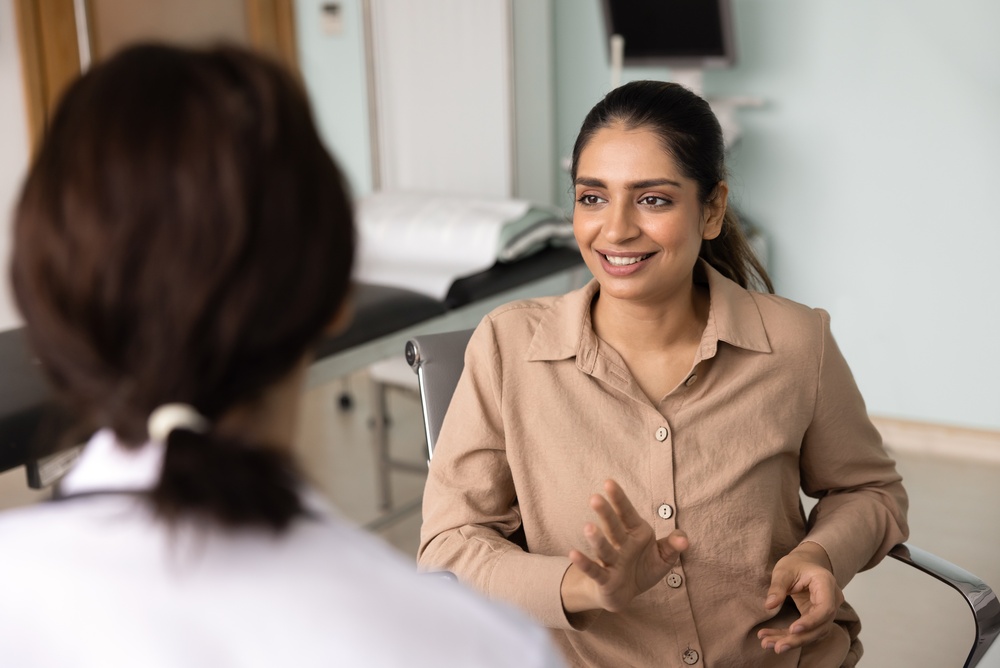 A woman in a beige shirt smiles and gestures with her hands while talking to another person, possibly a doctor, whose back is to the camera in a medical office. The mood appears friendly and engaged.